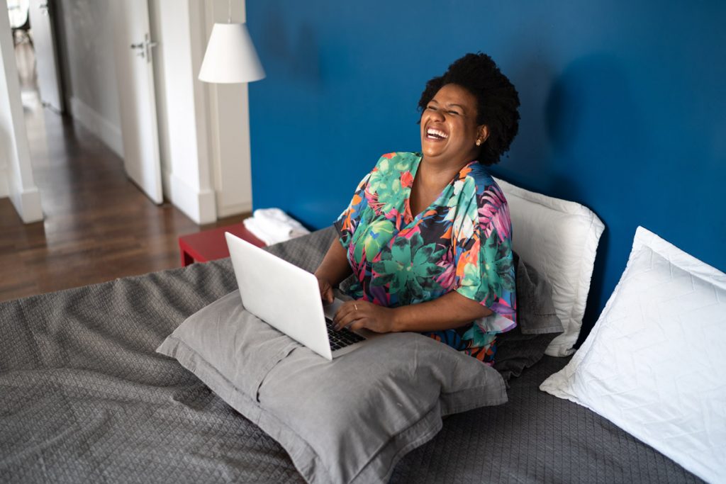Une femme rit alors qu'elle s'assoit sur son lit avec son ordinateur portable sur ses genoux.