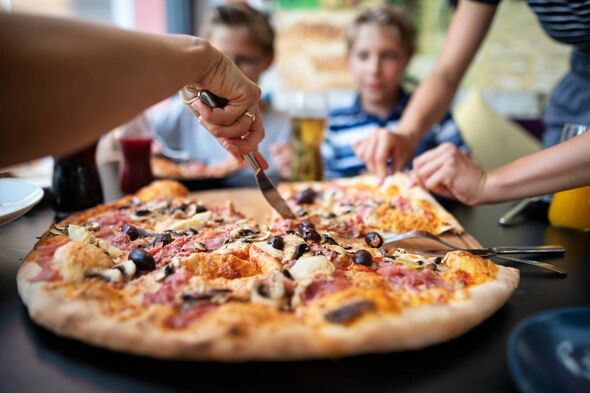 Plusieurs personnes sont rassemblées autour d'une grande pizza fraîchement cuite, avec de multiples garnitures dont divers légumes, viandes