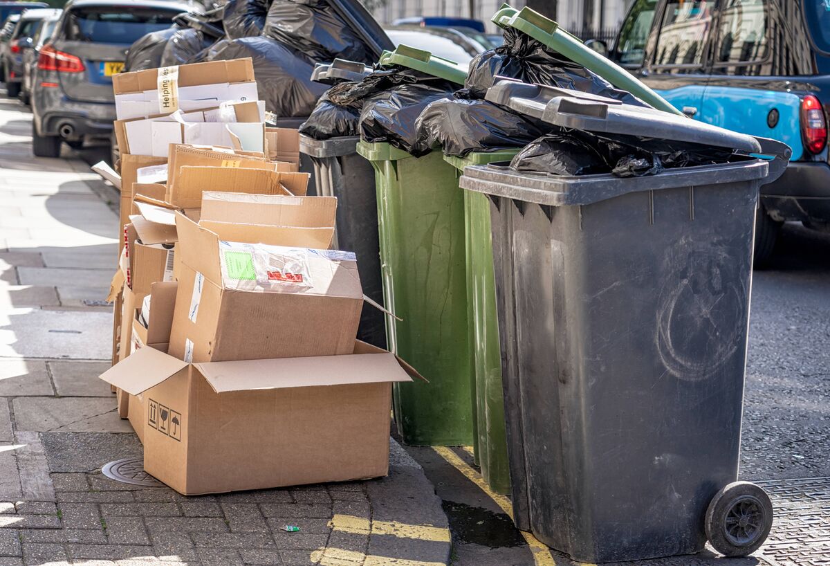 Un tas de carton dans une rue de Londres destiné au recyclage, à côté d'un groupe de poubelles roulantes en plastique débordantes.