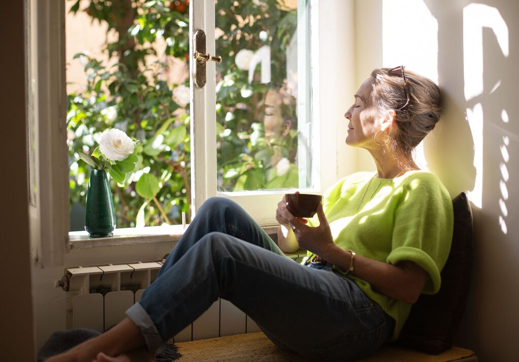 Femme se détendant dans un coin près de la fenêtre ouverte, profitant du soleil et d'une tasse de café Femme se détendant dans un coin près de la fenêtre ouverte, profitant du soleil et d'une tasse de café